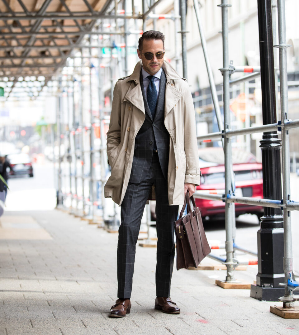 Khaki trench coat over a charcoal windowpane suit with blue tie and brown monk straps, styled on a city sidewalk