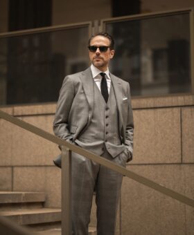 Grey three-piece wool suit with white shirt, black knit tie and white pocket square, worn by a model on outdoor steps