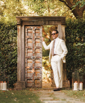 Ivory suit with white shirt, patterned tie and brown loafers, worn by a model by a garden gate