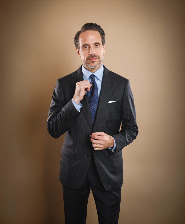 Charcoal striped two-button suit with blue striped shirt, navy dotted tie and white pocket square, worn by a model